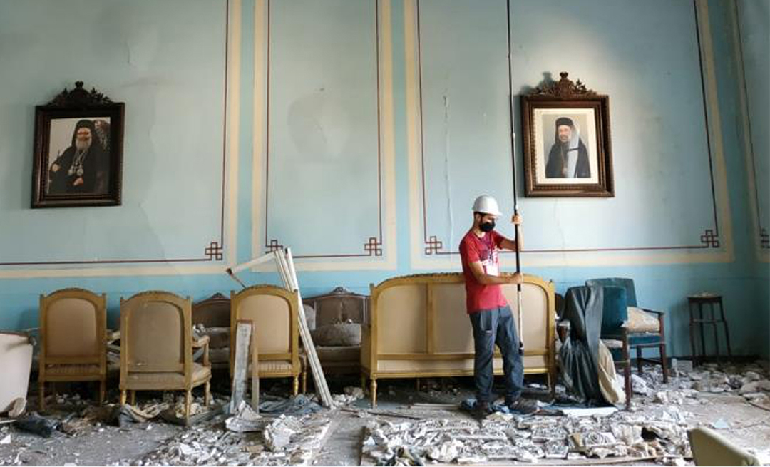 A person wearing a hard hat positions a camera on a tall stick in a blue room with clear damage on the floor and ceiling