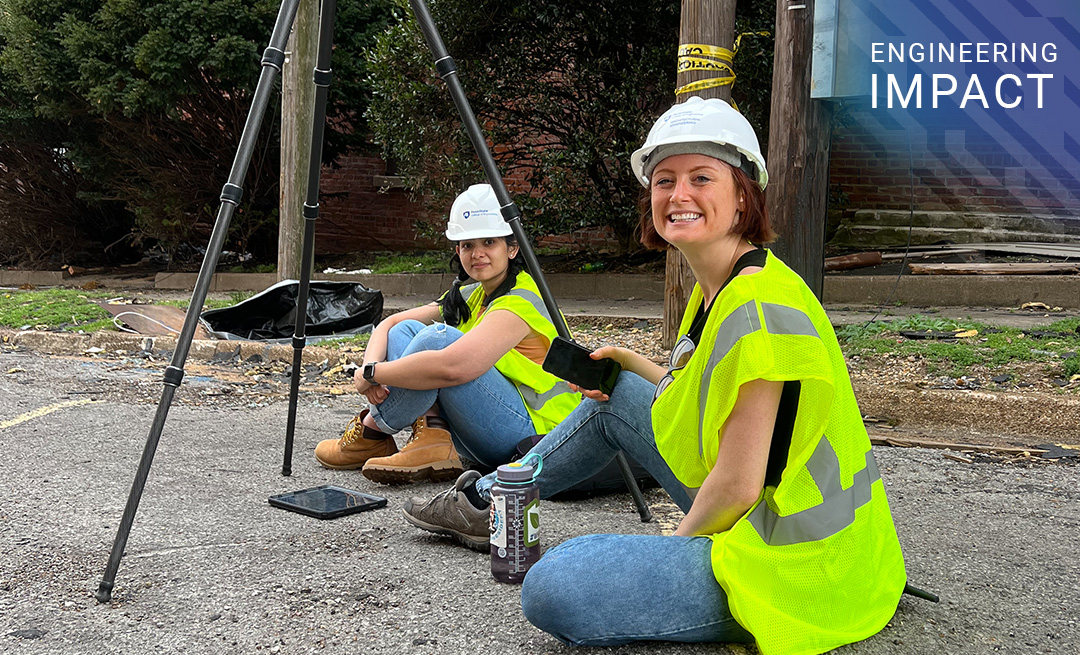 Two people wearing hard hats and safety vests sit on the ground 