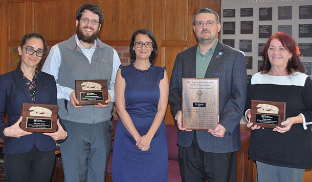 Two men and two women hold wooden plaques indoors next to another woman in the middle and smile at the camera.